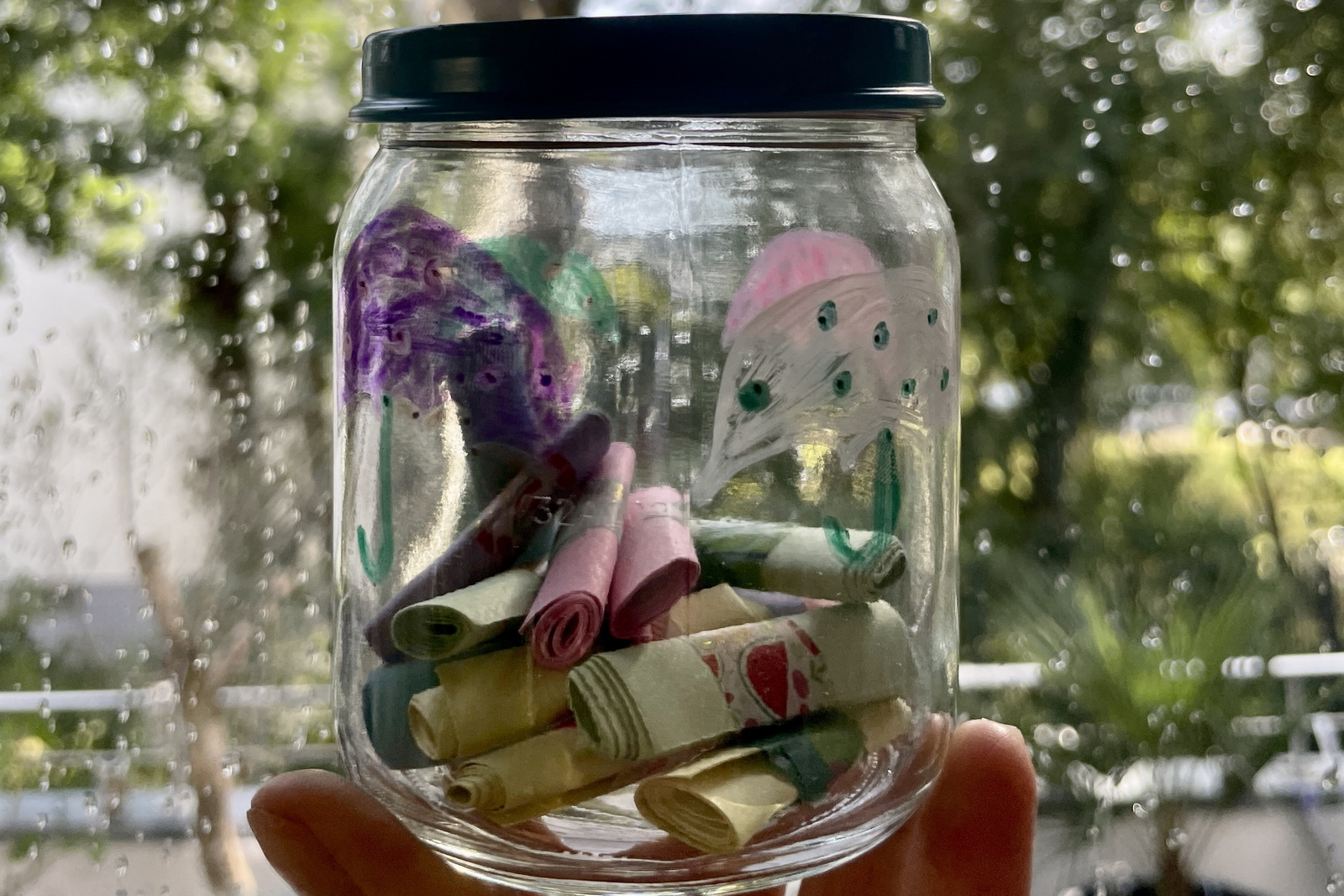 Bunte, handgeschriebene Zettel in einem Glas, gehalten vor einem regennassen Fenster mit unscharfem Hintergrund.