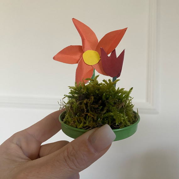 Bunte Papierblumen in einem kleinen grünen Topf mit Moos, gehalten von einer Hand.