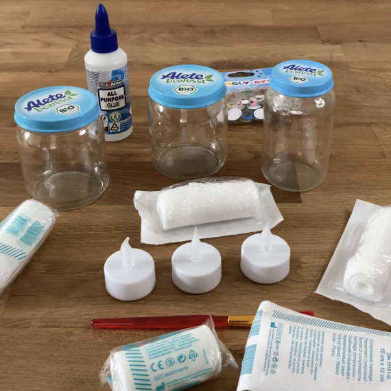 Materialien für DIY-Projekte: drei leere Gläser mit blauen Deckeln, Allzweckkleber, Kerzen und Bastelmaterialien auf Holzoberfläche.