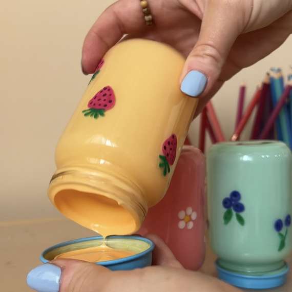 Hände gießen gelbe Flüssigkeit aus einem dekorierten Glas in einen blauen Behälter, umgeben von bunten Tassen.