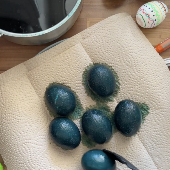 Blaue, lackierte Ostereier auf einem Küchenpapier, umgeben von einer Schüssel mit schwarzer Farbe und bunten Eierdekorationen.