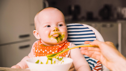 Baby mit grünem Brei im Gesicht, das aus einem Löffel gefüttert wird, sitzt in einem Hochstuhl mit gemusterter Rückenlehne.