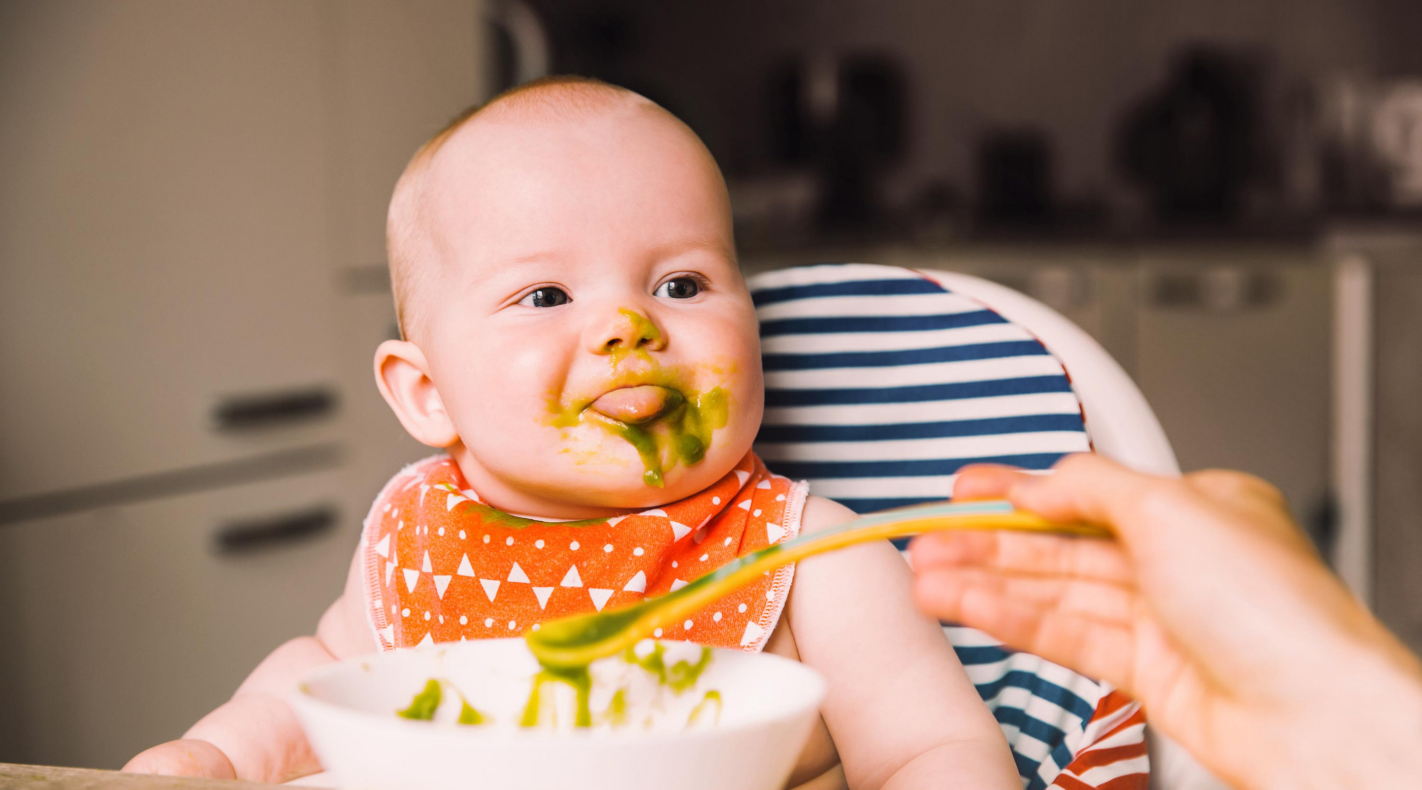 Baby mit grünem Brei im Gesicht, das aus einem Löffel gefüttert wird, sitzt in einem Hochstuhl mit gemusterter Rückenlehne.