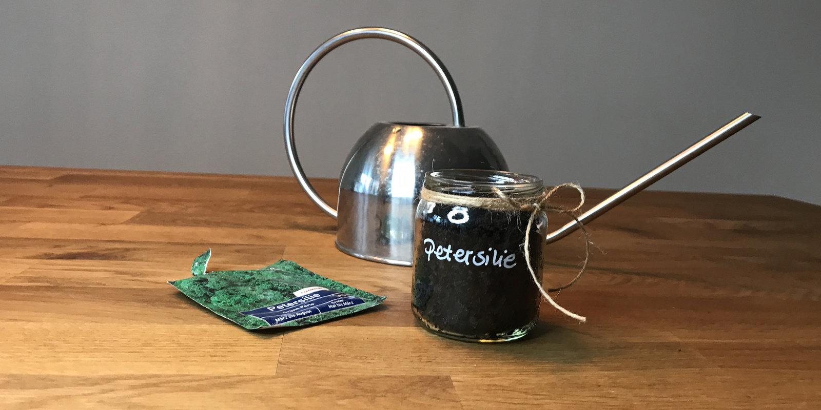 Gläserner Behälter mit Erde für Petersilie, daneben eine grüne Saatgutpackung und eine silberne Gießkanne auf Holzuntergrund.