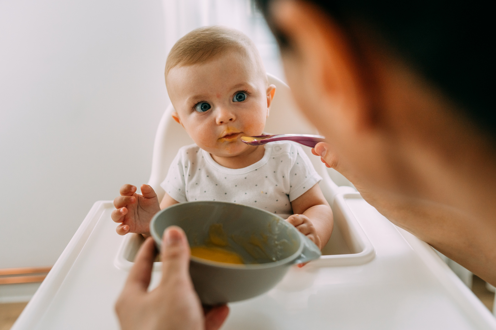 Babysitting beim Essen: Kleinkind in Hochstuhl schaut neugierig auf Löffel mit Brei aus grauer Schüssel.