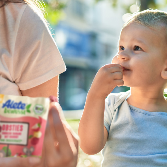 Ein Kleinkind genießt Alete Obsties im Freien, während eine erwachsene Person im Hintergrund die Packung festhält.