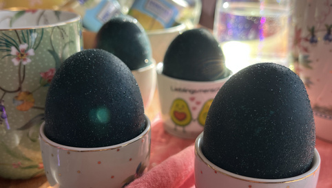 Dunkelblaue Ostereier in kleinen, dekorativen Eierbechern auf einer pinken Tischdecke, umgeben von Frühlingsblumen und dekorativen Elementen.