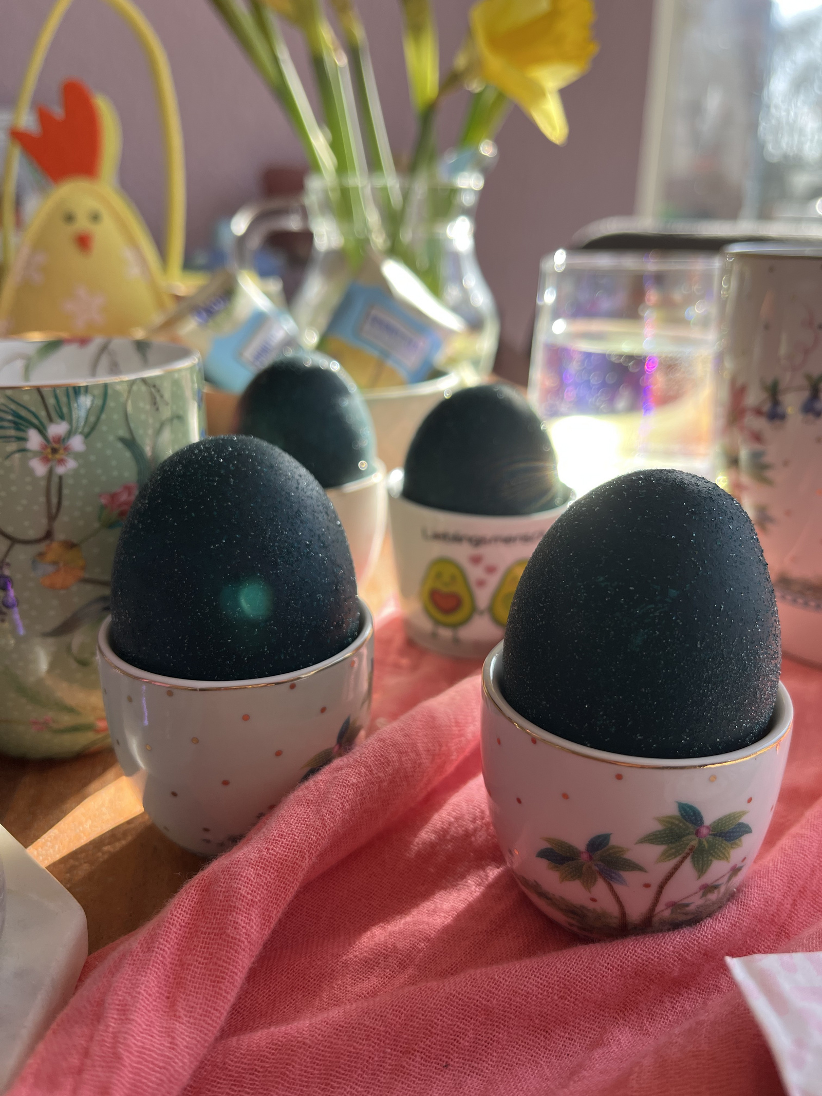 Dunkelblaue Ostereier in kleinen, dekorativen Eierbechern auf einer pinken Tischdecke, umgeben von Frühlingsblumen und dekorativen Elementen.