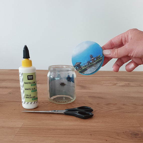 Bastelmaterialien auf einem Tisch: eine Flasche Bastelkleber, eine Schere und ein rundes Bild eines Strandmotivs, während eine Person das Bild hält.