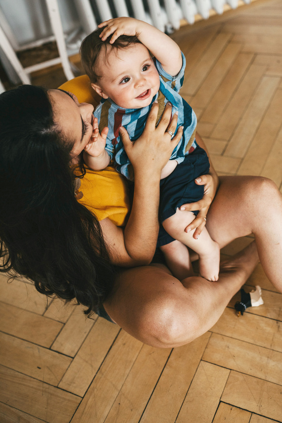 Mutter und Baby lachen gemeinsam auf dem Boden, während das Baby mit einer Hand den Kopf hält und fröhlich wirkt.