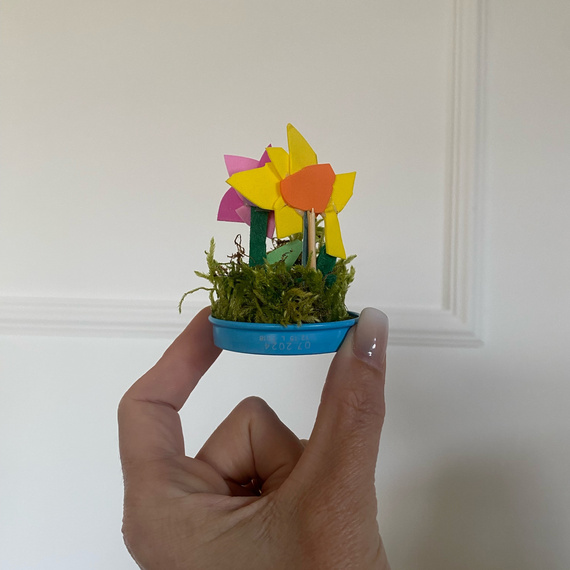 Bunte Papierblumen in einem Topf mit Moos, gehalten von einer Hand, auf einem einfarbigen Hintergrund.