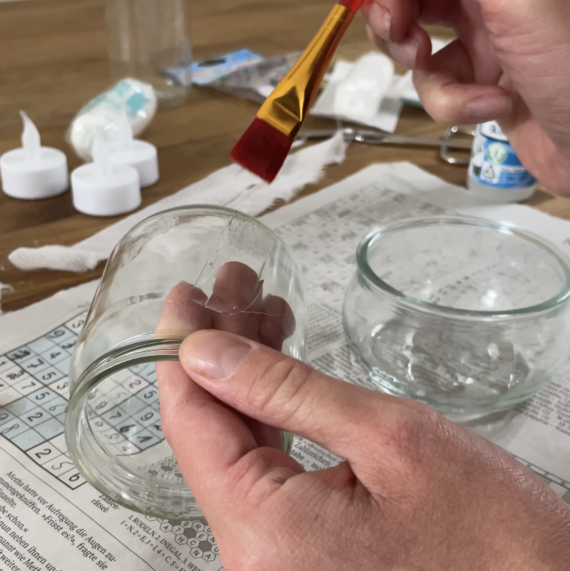 Händchen mit Pinsel bemalt ein Glasgefäß, während sich weitere Bastelmaterialien und Zeitungsartikel auf dem Tisch befinden.