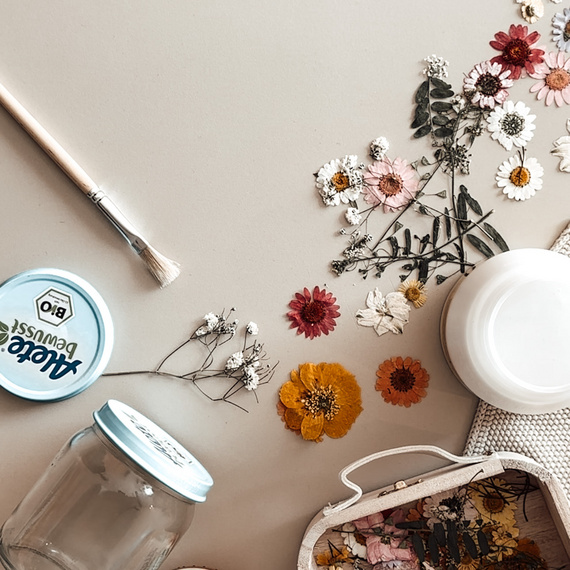 Bunte, getrocknete Blumen und Bastelmaterialien auf einem Tisch, umgeben von einem Pinsel, einem leeren Glas und einer dekorativen Box.
