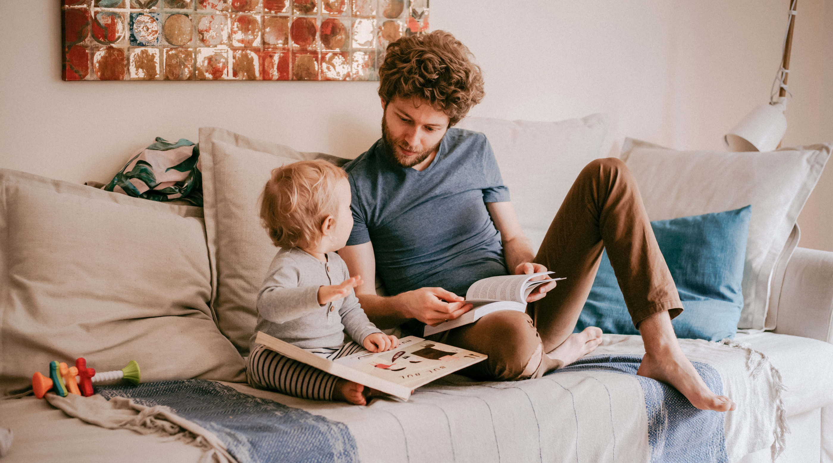 Vater und Kleinkind sitzen entspannt auf einem Sofa und lesen gemeinsam Bücher. Bunte Spielzeuge liegen auf dem Sofa.