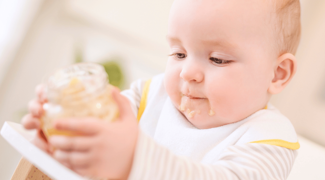 Ein Baby mit einem Lätzchen hält ein Glas mit einer cremigen Speise und hat dabei eine schmutzige Mundpartie.