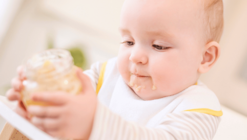 Ein Baby mit einem Lätzchen hält ein Glas mit einer cremigen Speise und hat dabei eine schmutzige Mundpartie.
