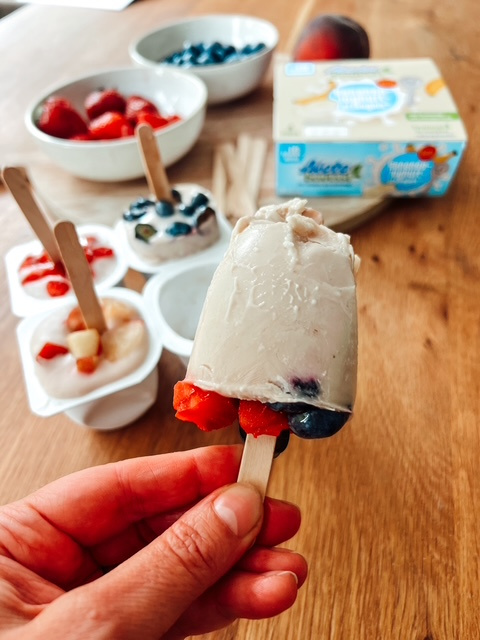 Fruchtiges Jogurt-Eis am Stiel mit Erdbeeren und Heidelbeeren, umgeben von frischen Beeren und Joghurtbechern auf einem Holztisch.