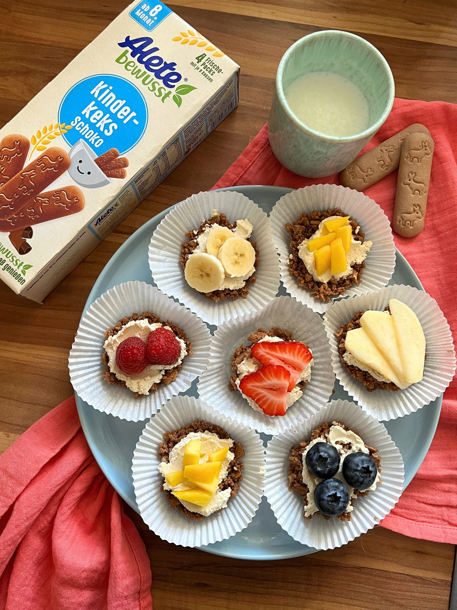 Bunte Kekstörtchen mit einer Schicht Quark und verschiedenen Früchten, serviert auf einem Teller mit Alete Kinder-Keksen und einem Glas Milch.