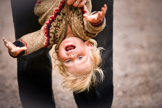 Lachendes Kind mit blonden Haaren, das auf dem Kopf hängend von einer erwachsenen Person gehalten wird, draußen auf unebenem Untergrund.