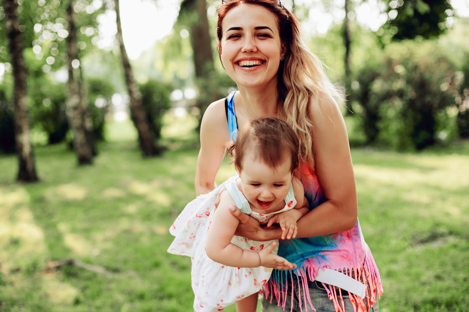 Lächelnde Mutter trägt fröhliches Kleinkind auf einer Wiese, umgeben von Bäumen, in einer sonnigen Umgebung.