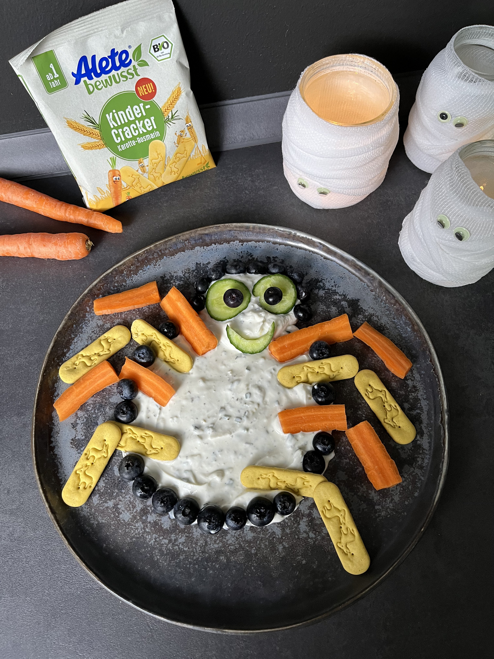 Bunte Spinne aus Quark, Karotten, Gurken, blauen Beeren und Alete Kinder-Crackern auf einem schwarzen Hintergrund, dekoriert mit Spuklichtern.