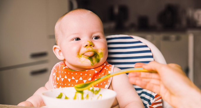Baby mit grünem Brei im Gesicht, das aus einem Löffel gefüttert wird, sitzt in einem Hochstuhl mit gemusterter Rückenlehne.