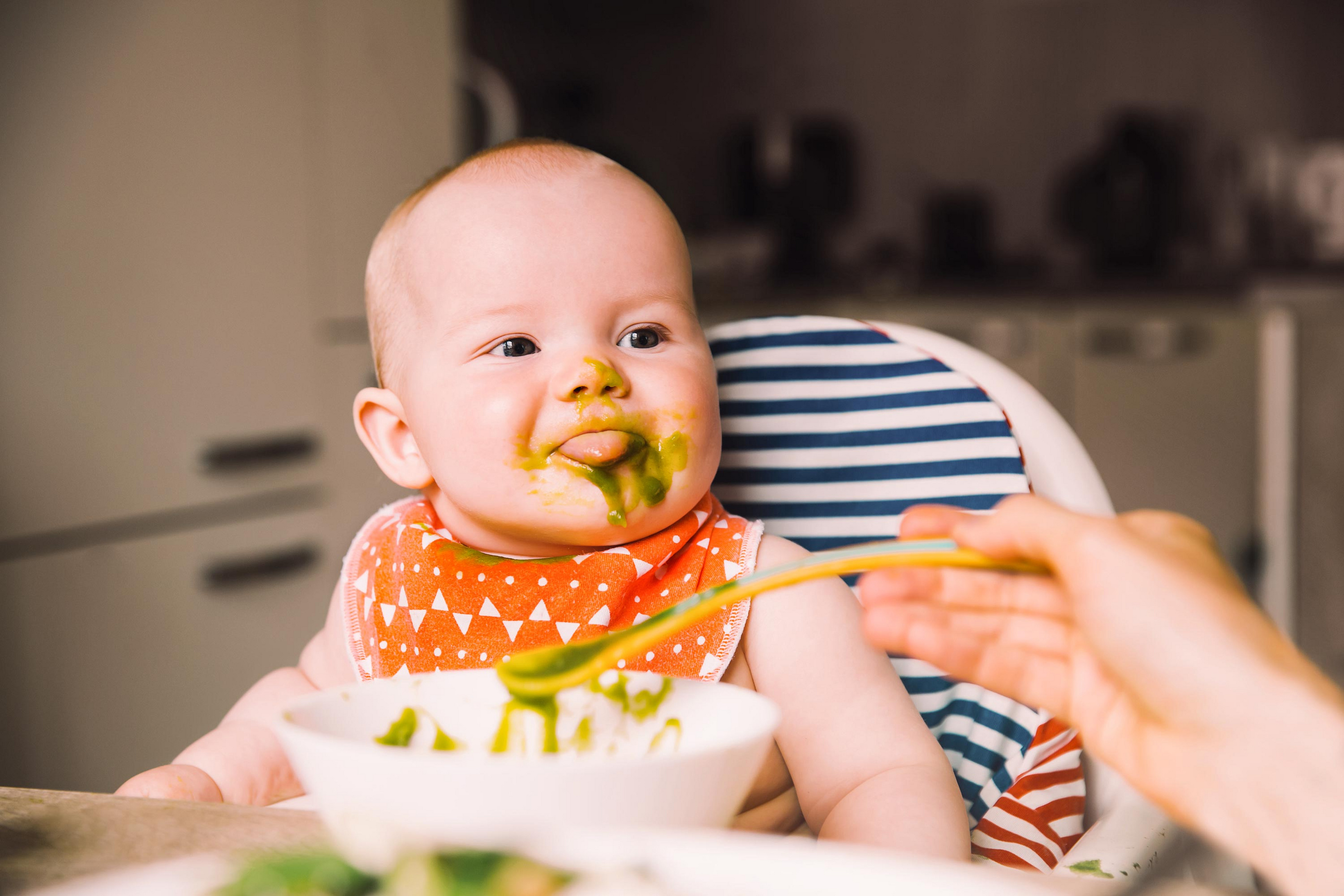Baby mit grünem Brei im Gesicht, das aus einem Löffel gefüttert wird, sitzt in einem Hochstuhl mit gemusterter Rückenlehne.