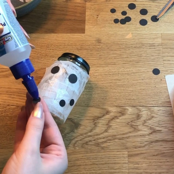 Hand, die einen Kleber auf einen Glasbehälter aufträgt, der mit schwarzem Papier dekoriert wird. Bastelmaterialien und Werkzeug auf einem Holztisch.