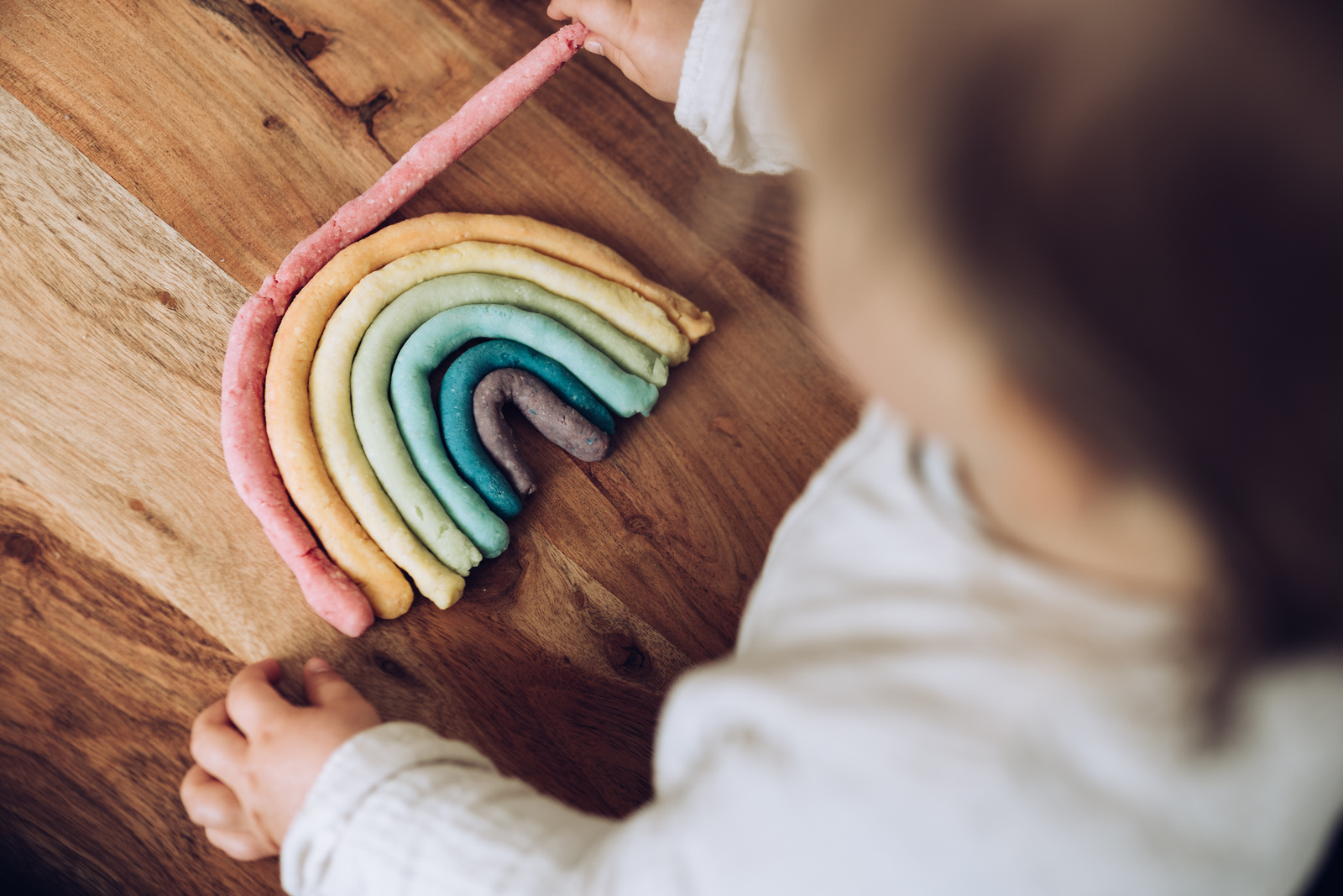 Bunte Knetebogen in Regenbogenform auf einem Holztisch, während ein Kind damit spielt.