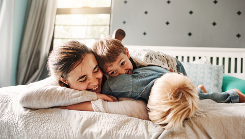 Lächelnde Mutter mit zwei Kindern, die auf einem Bett spielen und kuscheln, in einem hellen und einladenden Raum.