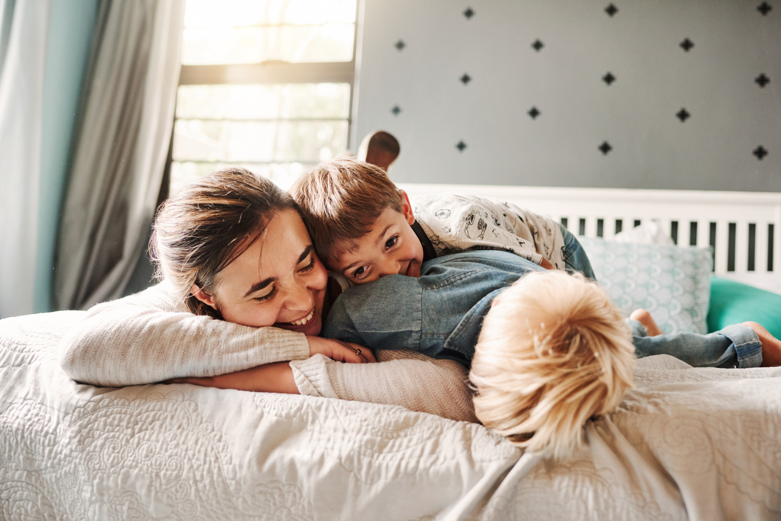Lächelnde Mutter mit zwei Kindern, die auf einem Bett spielen und kuscheln, in einem hellen und einladenden Raum.
