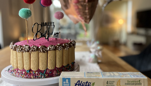 Bunter Geburtstagskuchen mit Luftballons und Kinderkeks-Packung auf einem Tisch. Der Kuchen ist mit bunten Streuseln und einem „Happy Birthday“-Schriftzug dekoriert.