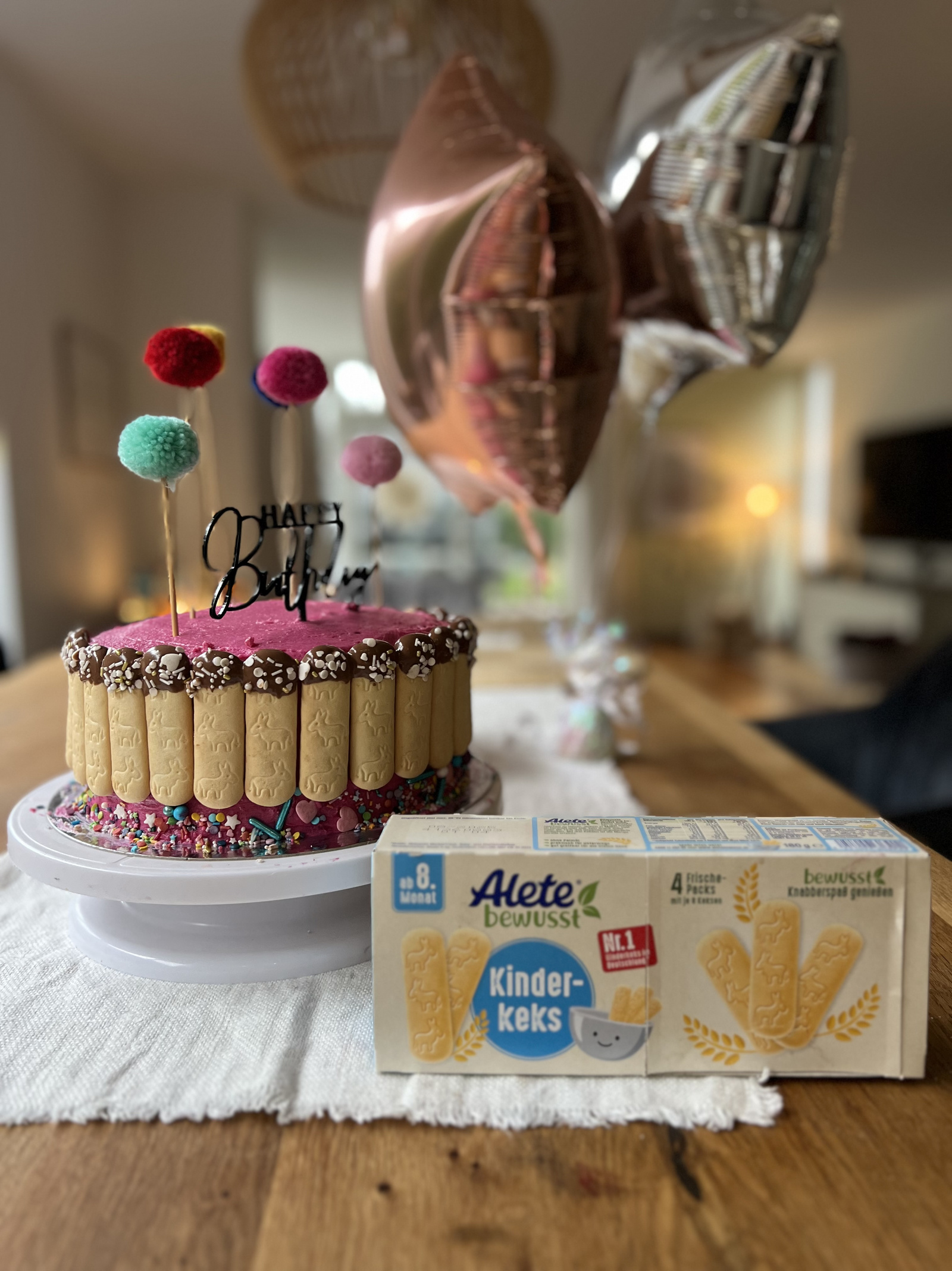 Bunter Geburtstagskuchen mit Luftballons und Kinderkeks-Packung auf einem Tisch. Der Kuchen ist mit bunten Streuseln und einem „Happy Birthday“-Schriftzug dekoriert.
