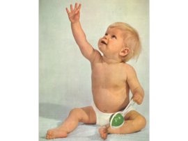 Ein Kleinkind mit blonden Haaren sitzt auf dem Boden und hebt den Arm, während es ein grünes Spielzeug betrachtet.