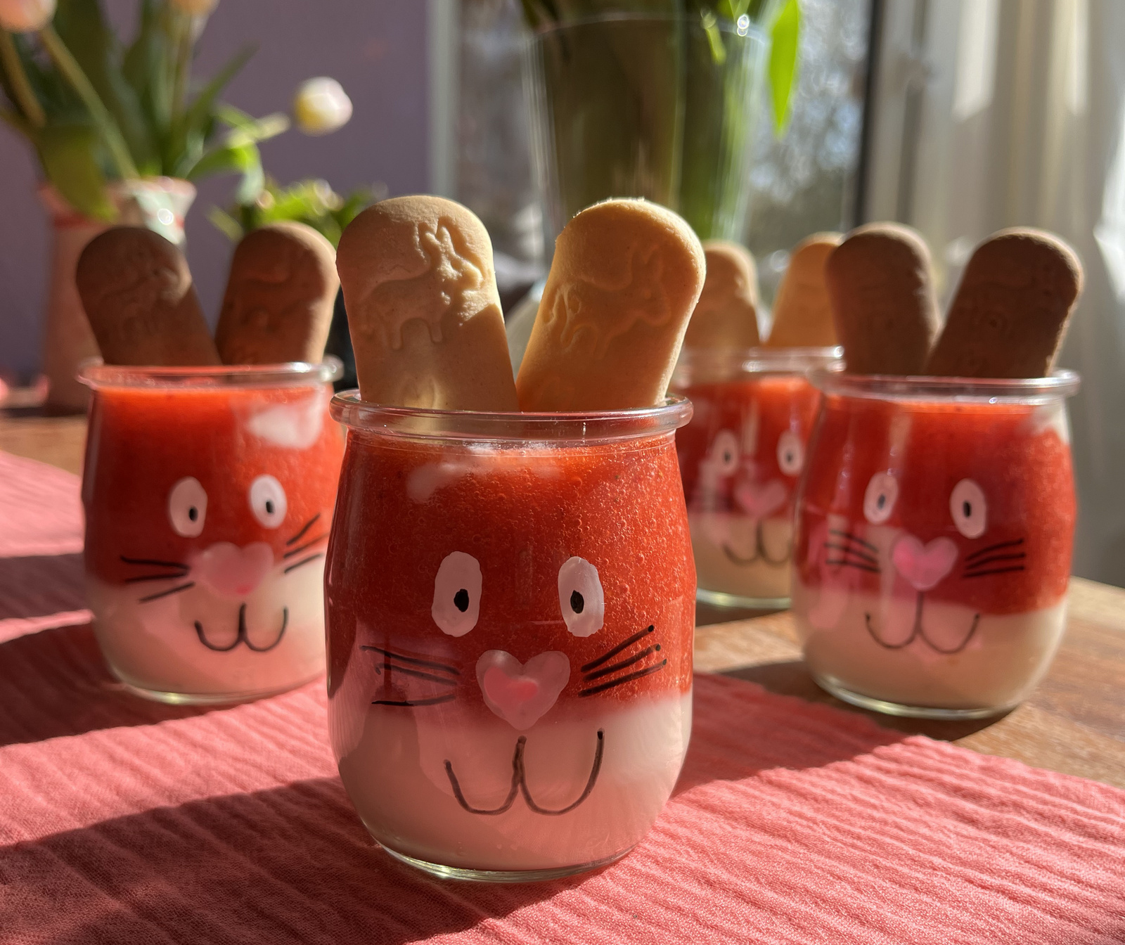 Süße Dessertgläser in Hasenform mit Erdbeer-Topping und Keksohren auf einem rosa Tischtuch, umgeben von frischen Tulpen.