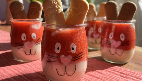 Süße Dessertgläser in Hasenform mit Erdbeer-Topping und Keksohren auf einem rosa Tischtuch, umgeben von frischen Tulpen.