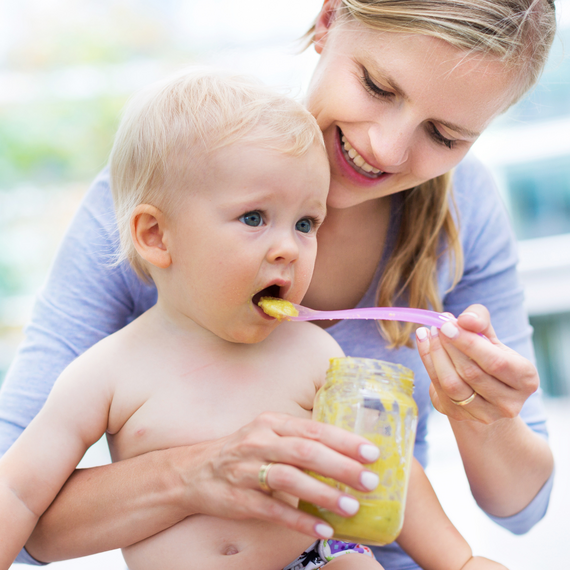 Mutter füttert ihr Kleinkind mit einer Löffelportion Babybrei im Freien, umgeben von unscharfen, grünen Bäumen.