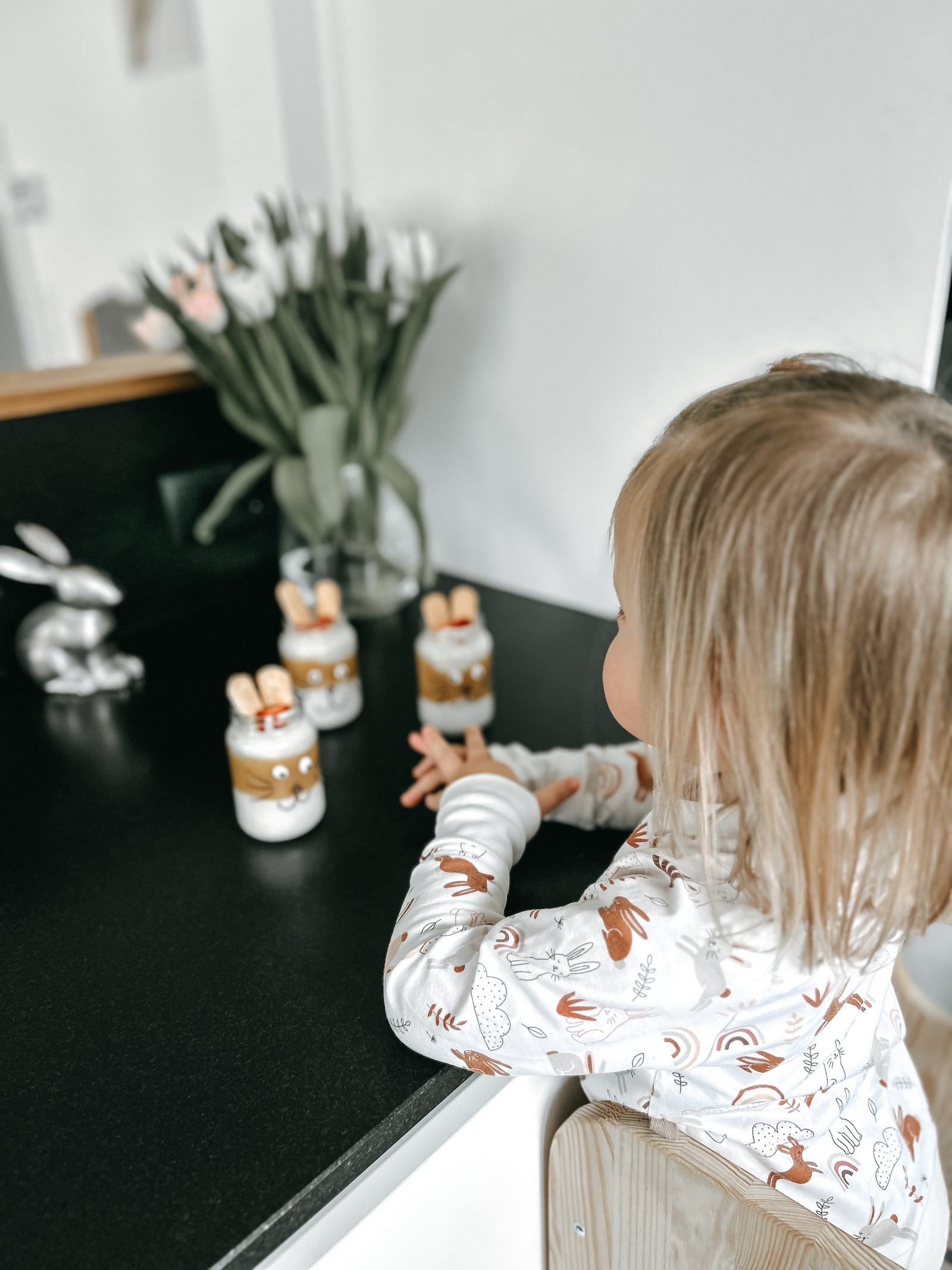 Kleines Kind in einem weißen Oberteil mit Hasenmotiven, sitzt an einem Tisch und schaut auf dekorierte Gläser mit Süßigkeiten und einer Vase mit Tulpen.