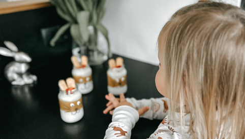 Kleines Kind in einem weißen Oberteil mit Hasenmotiven, sitzt an einem Tisch und schaut auf dekorierte Gläser mit Süßigkeiten und einer Vase mit Tulpen.