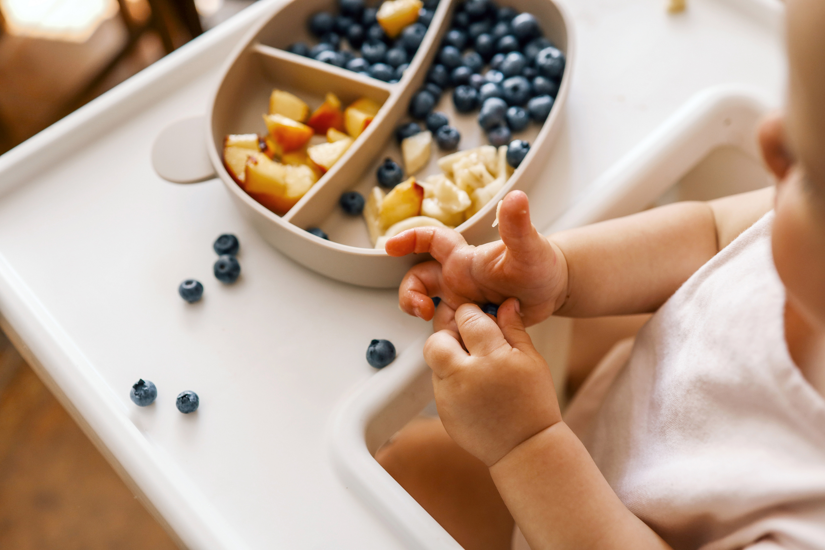 Kleinkind sitzt am Hochstuhl und greift nach Heidelbeeren aus einem Teller mit geschnittenem Obst.