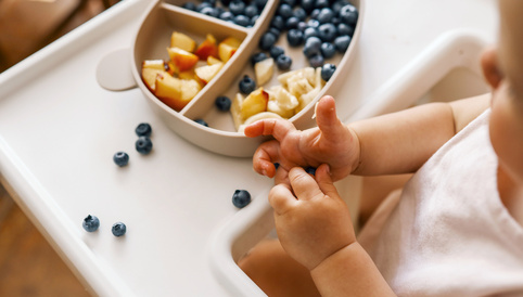 Kleinkind sitzt am Hochstuhl und greift nach Heidelbeeren aus einem Teller mit geschnittenem Obst.