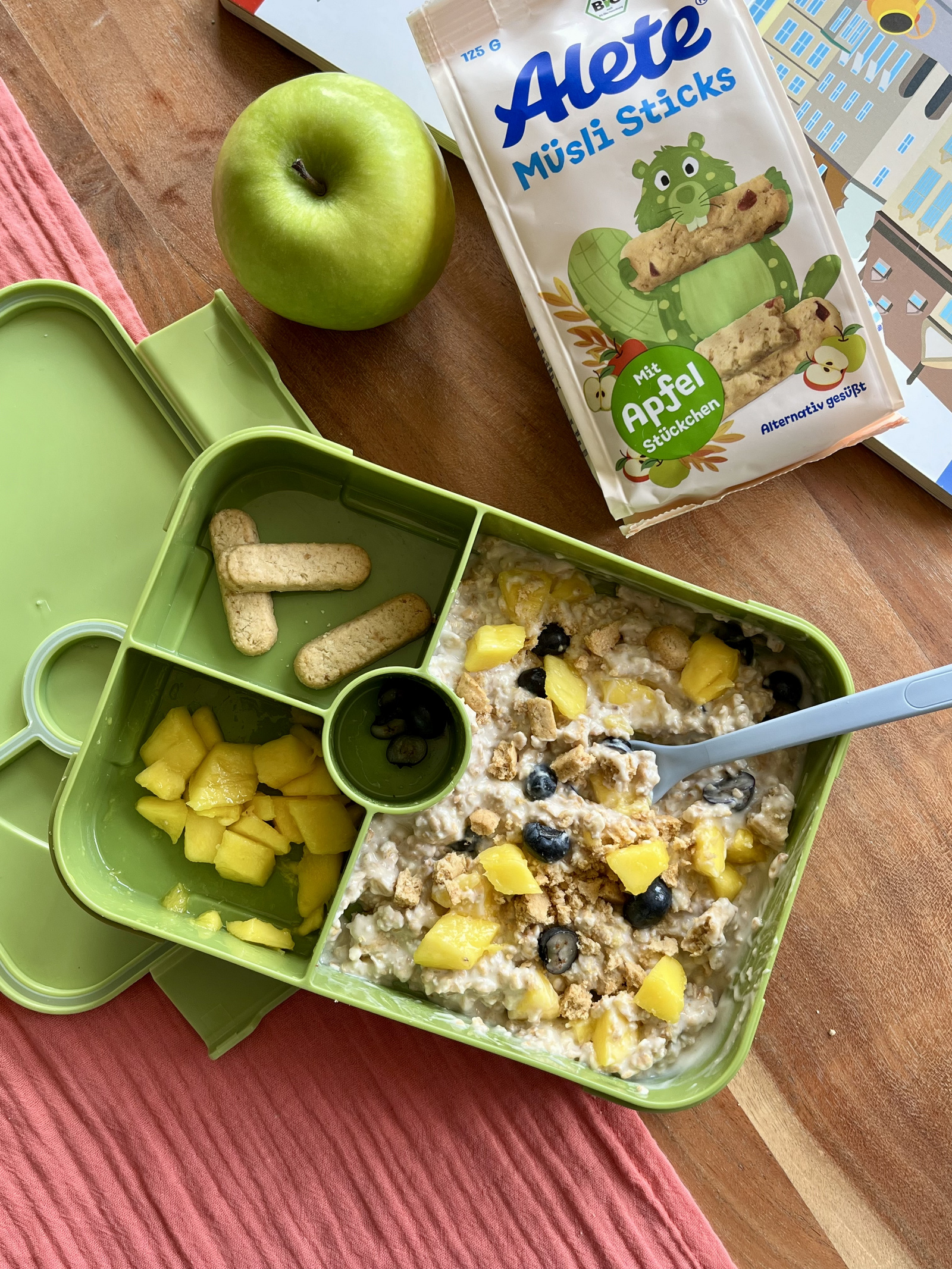 Gesunde Lunchbox mit OvernightOats frischen Mango- und Heidelbeerstücken, Müsliriegeln und einem grünen Apfel.
