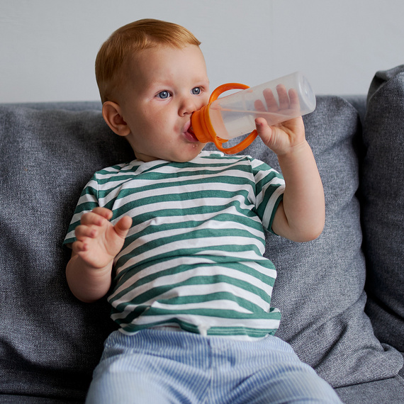Kleinkind sitzt auf grauem Sofa und trinkt aus einer Trinklern-Flasche