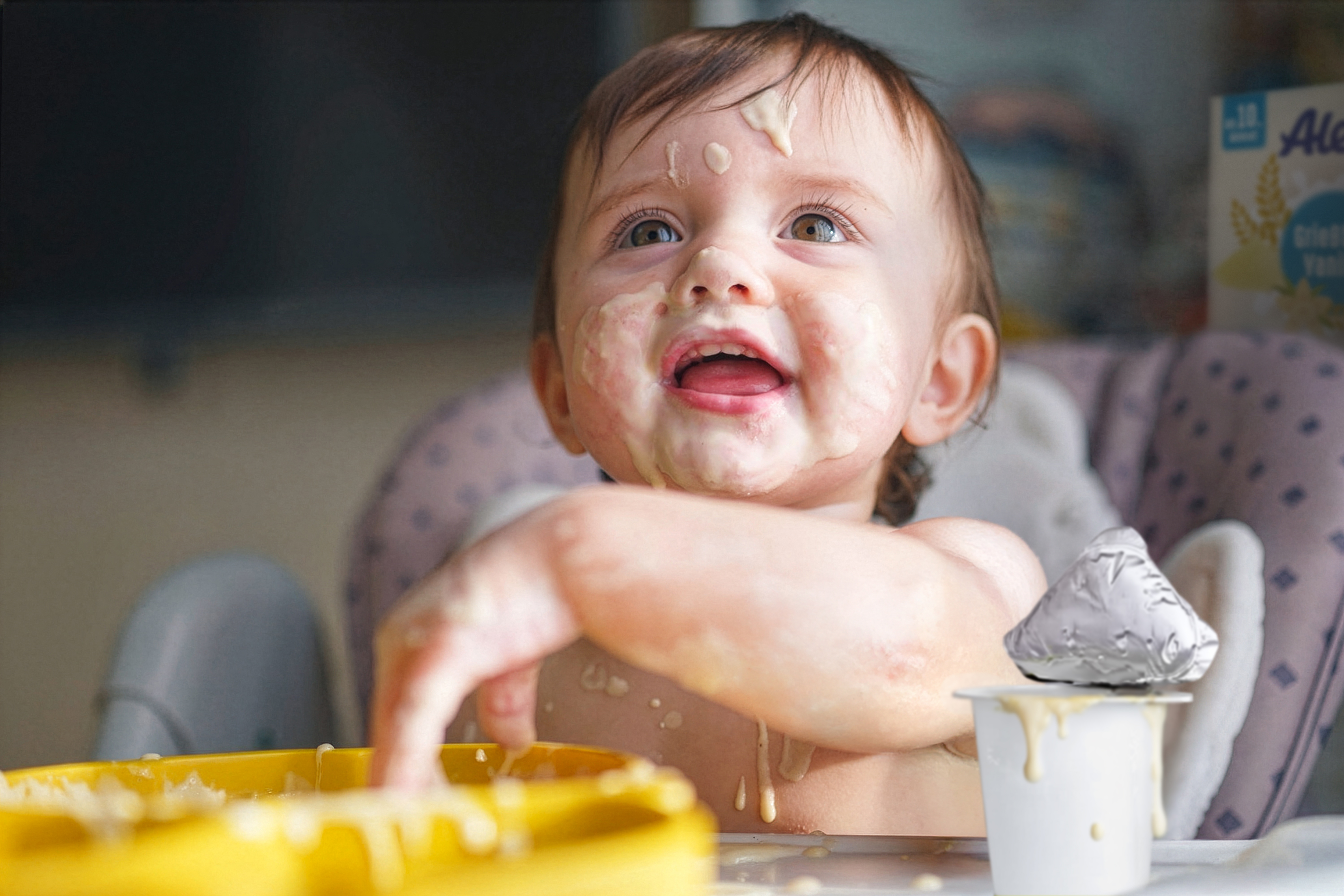 Lächelndes Baby mit Essensreste im Gesicht, das aus einem gelben Schälchen isst, neben einem Glas Alete Abendbrei.
