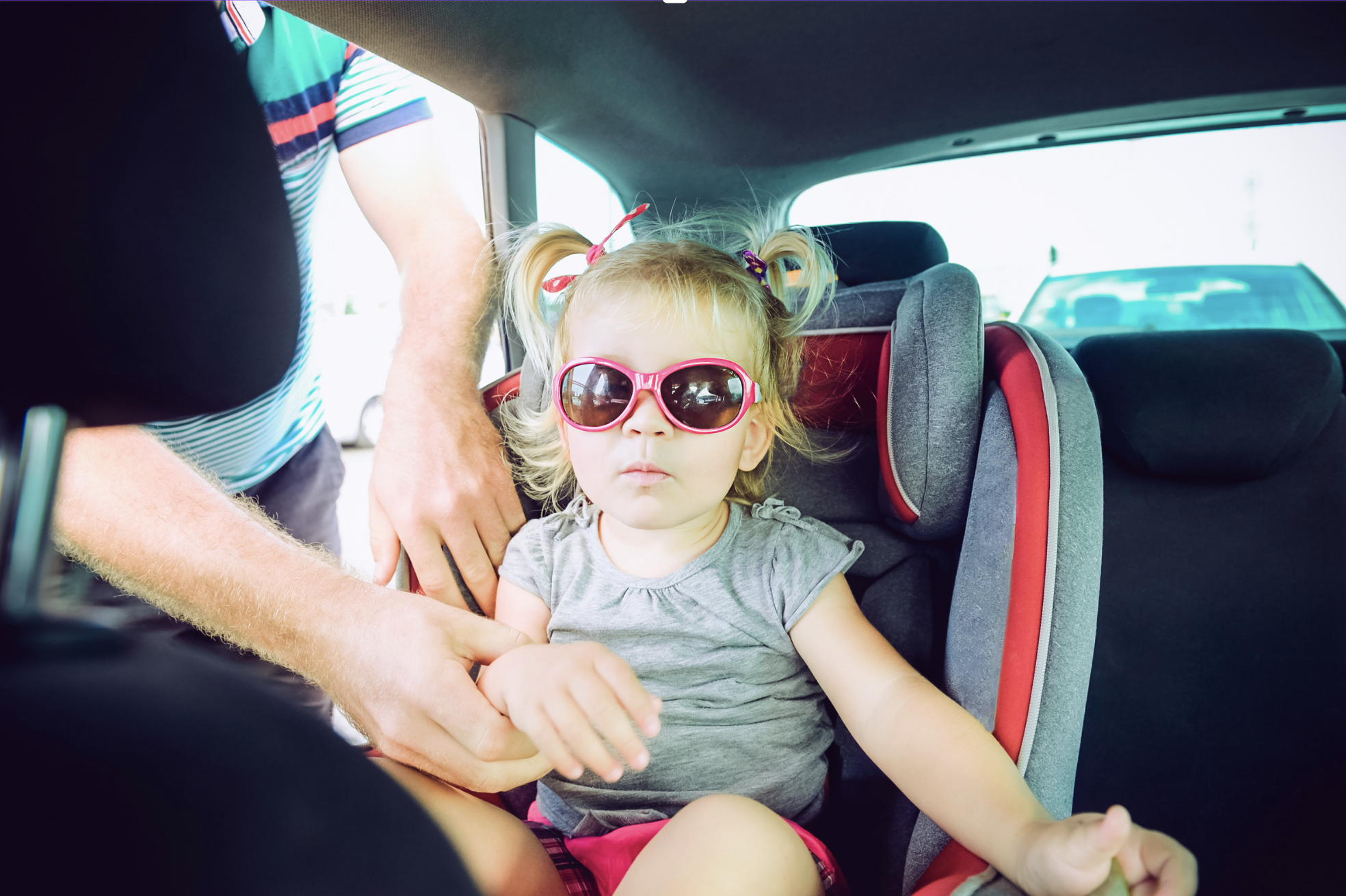 Ein kleines Mädchen in Sonnenbrille sitzt in einem Autositz, während eine Hand sie anschnallt.