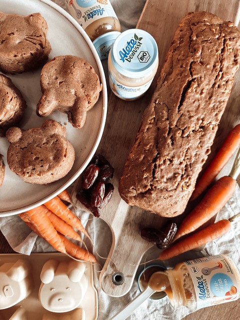 Karottenkuchen in Kasten- und Hasenform liegt auf Holzbrett, daneben Karotten und Alete-Gläschen